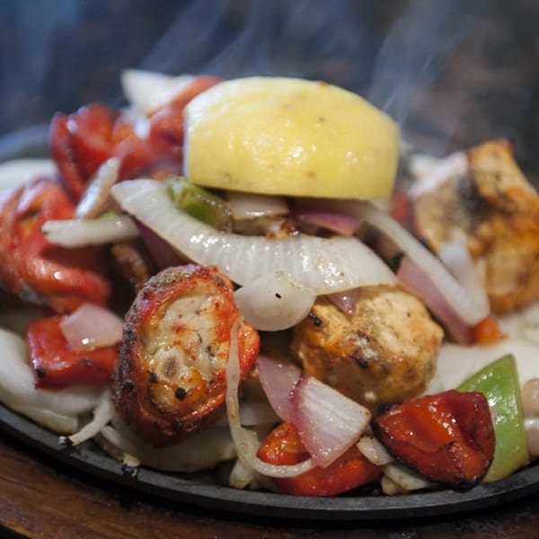 Sizzling chicken tikka with onions, peppers, and a lemon wedge on a dark plate.