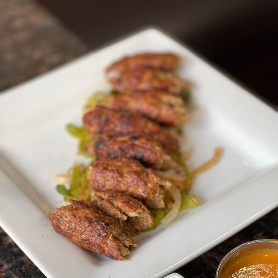 Seven grilled kebab pieces on a bed of green and white onions, with a dipping sauce.