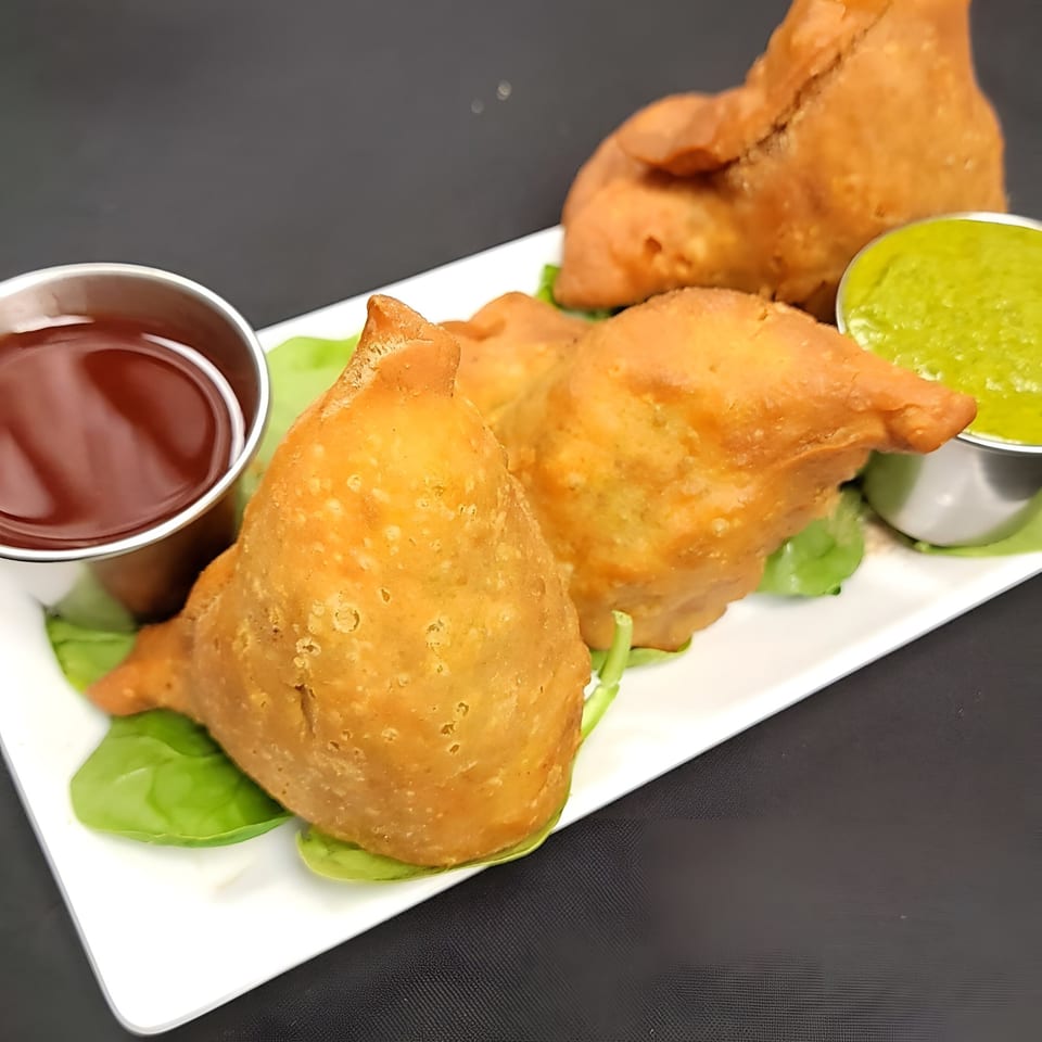 Golden-brown samosas on a white plate with green chutney and red sauce, from Spice Hub.